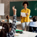 Happy African American teacher holding a class and pointing at student with raised arm who wants to answer the question.