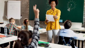Happy African American teacher holding a class and pointing at student with raised arm who wants to answer the question.
